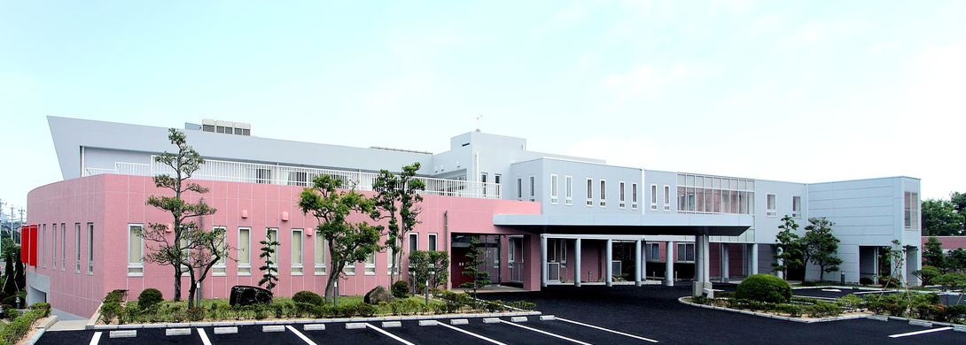 ピンク色と白を基調とした現代的な介護施設の外観。広い駐車場と周辺植栽が見える。