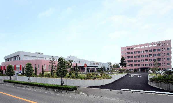 介護施設の建物外観。ピンク色の壁と白い建物が特徴的。駐車場と緑地がある。
